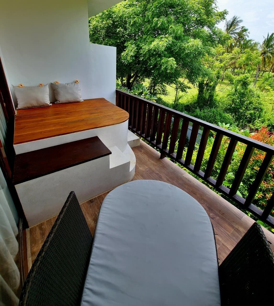 A balcony with a wooden bench and a white cushioned seating area, overlooking a lush garden.