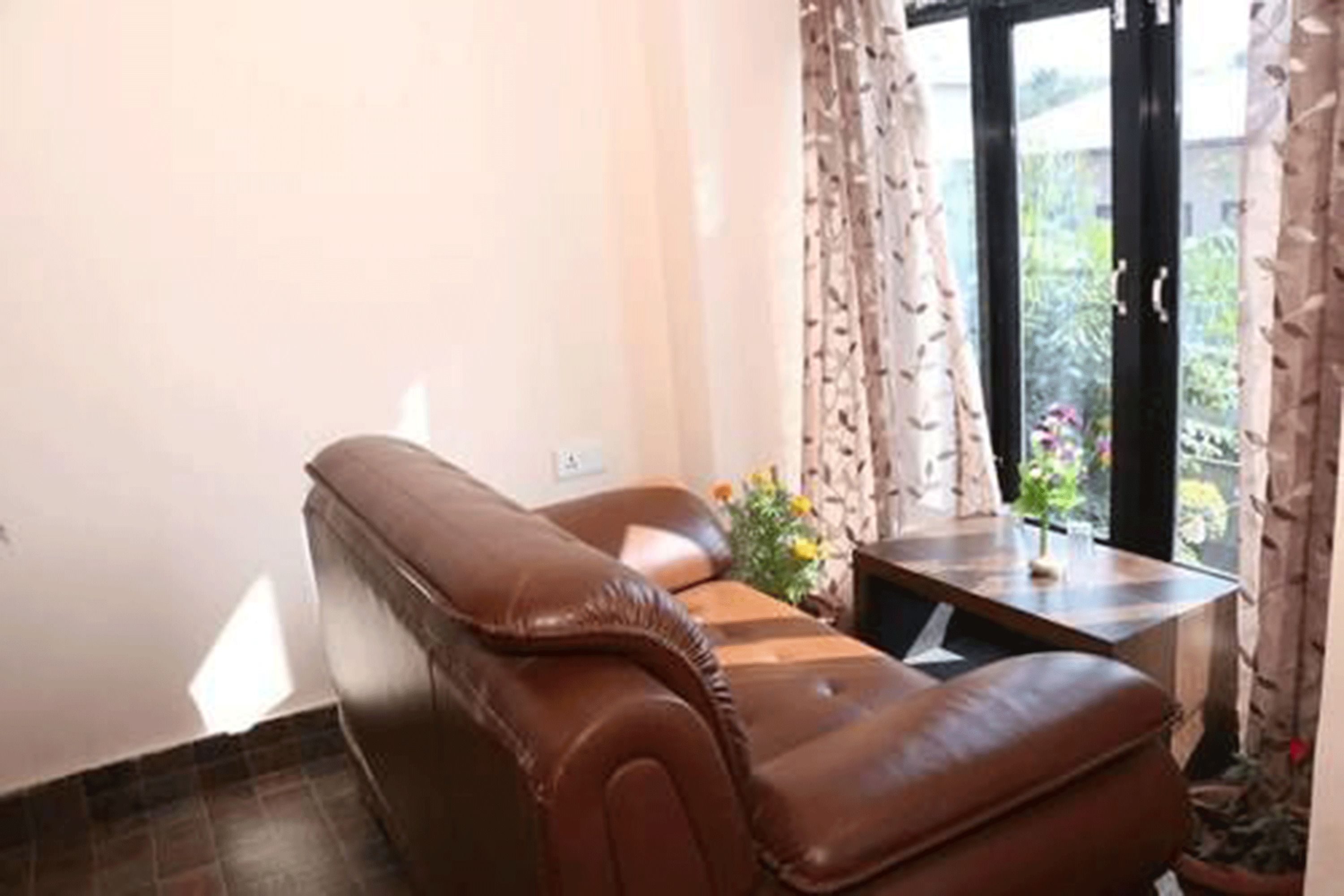 Un sofá de cuero marrón en una habitación con ventana, cortinas y una pequeña mesa con un florero.