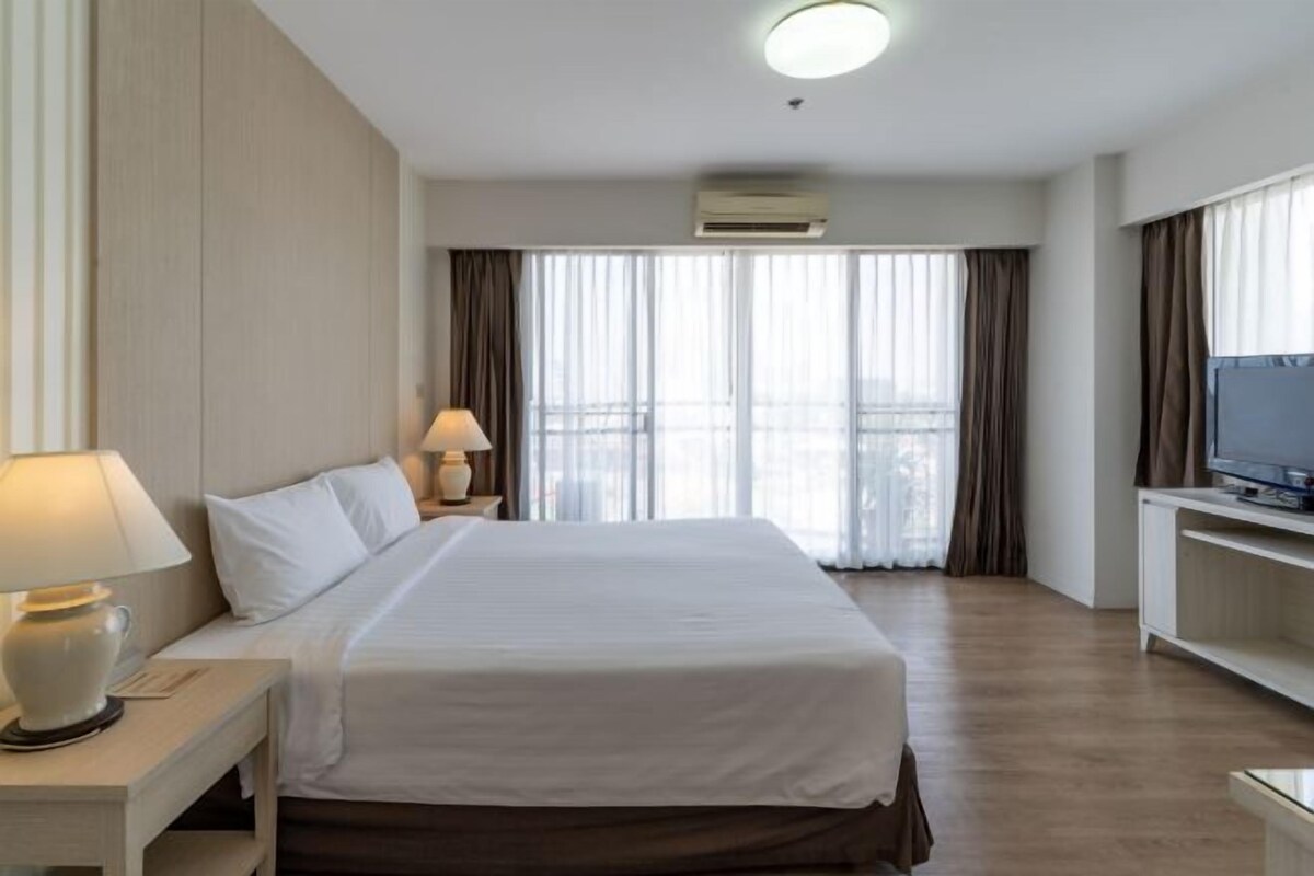 Ein Hotelzimmer mit einem großen Bett, einem Fernseher auf einem Tisch, einem Nachttisch mit Lampe und einem Fenster mit Vorhängen.