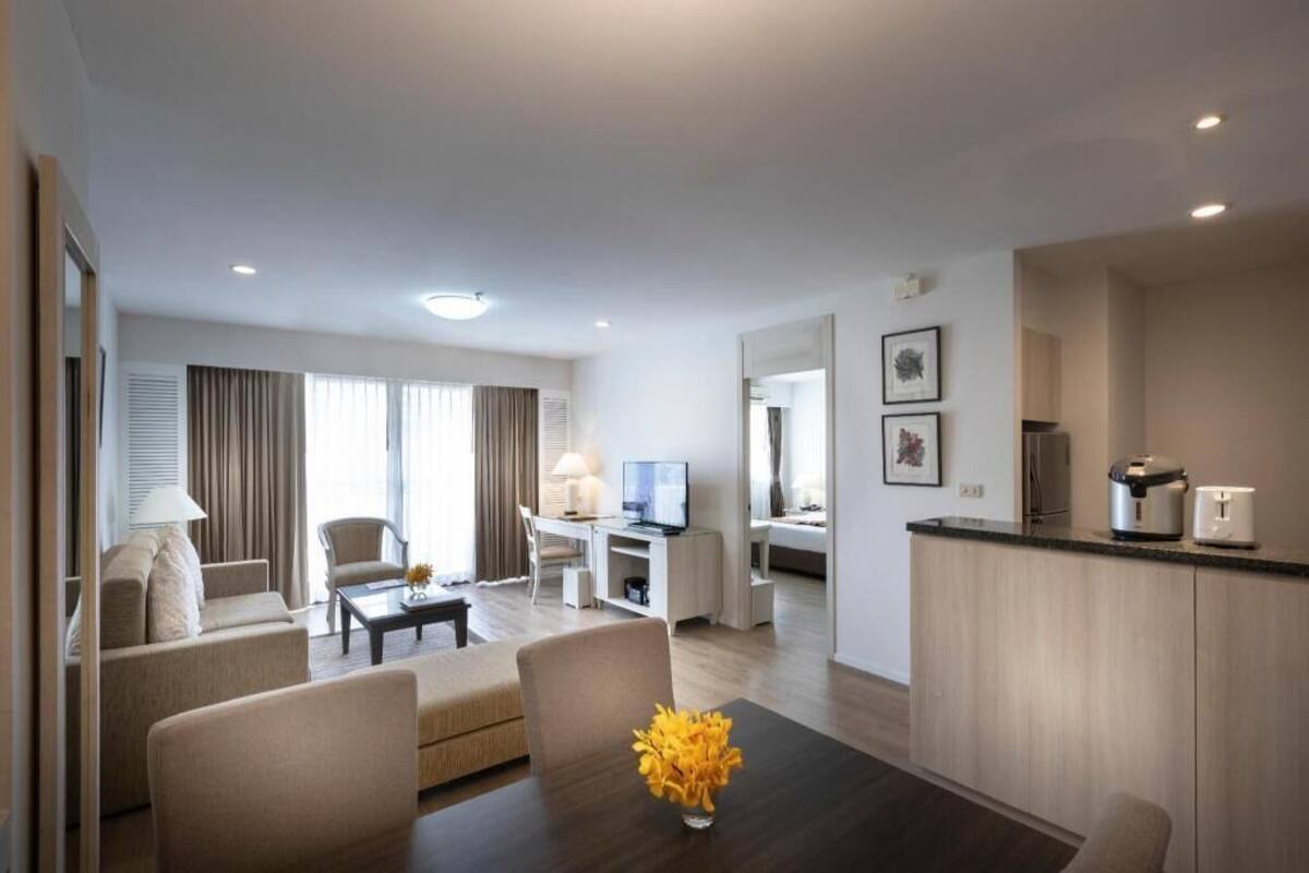 Ein modernes Hotelzimmer mit Wohnbereich, einem Esstisch mit Blumenvase, einer Küchenzeile und einem Fernseher.