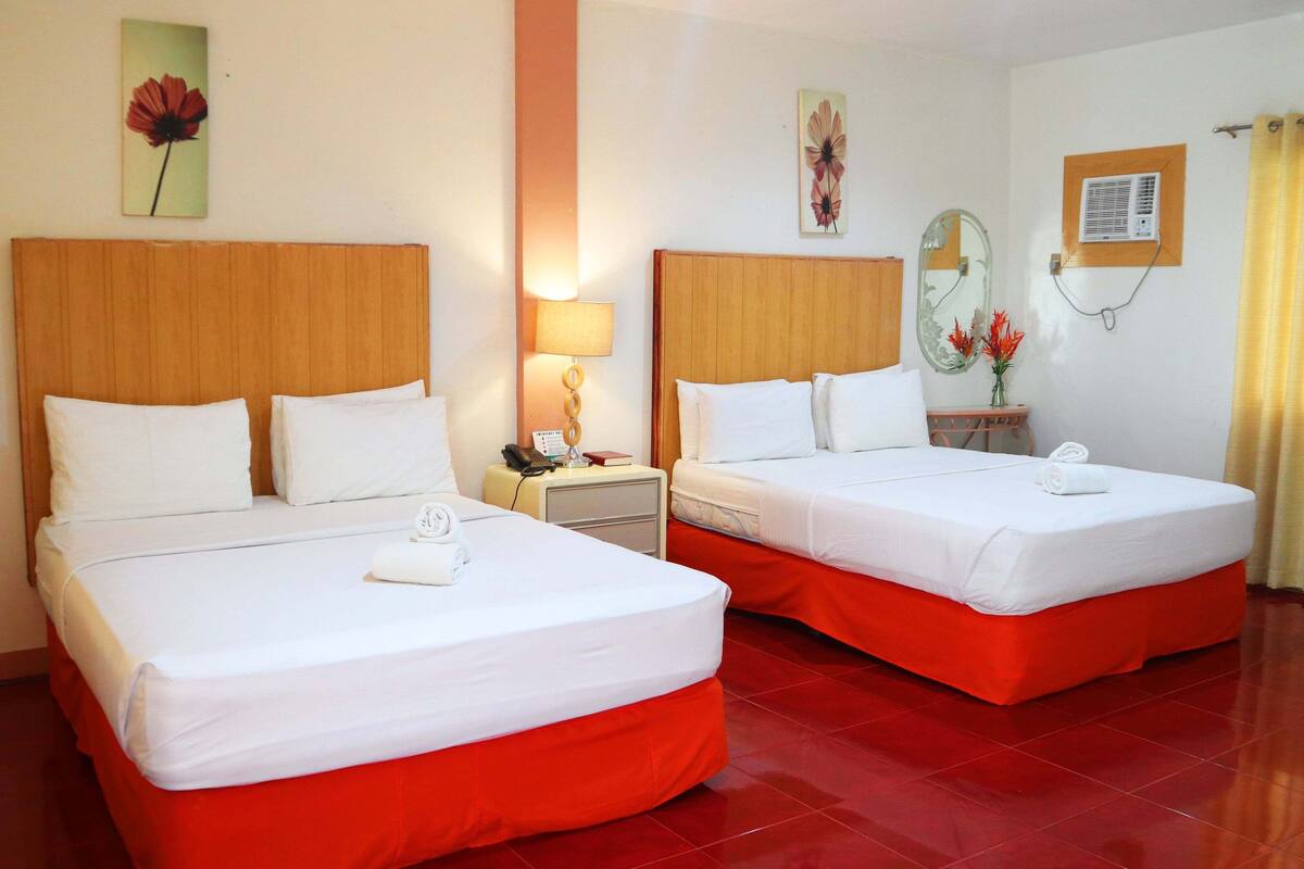 A hotel room with two beds, each with white linens and red bed skirts. There is a nightstand with a lamp, a telephone, and a small table with a vase of flowers. The room has a wooden headboard, a wall-mounted air conditioning unit, and artwork above the beds.