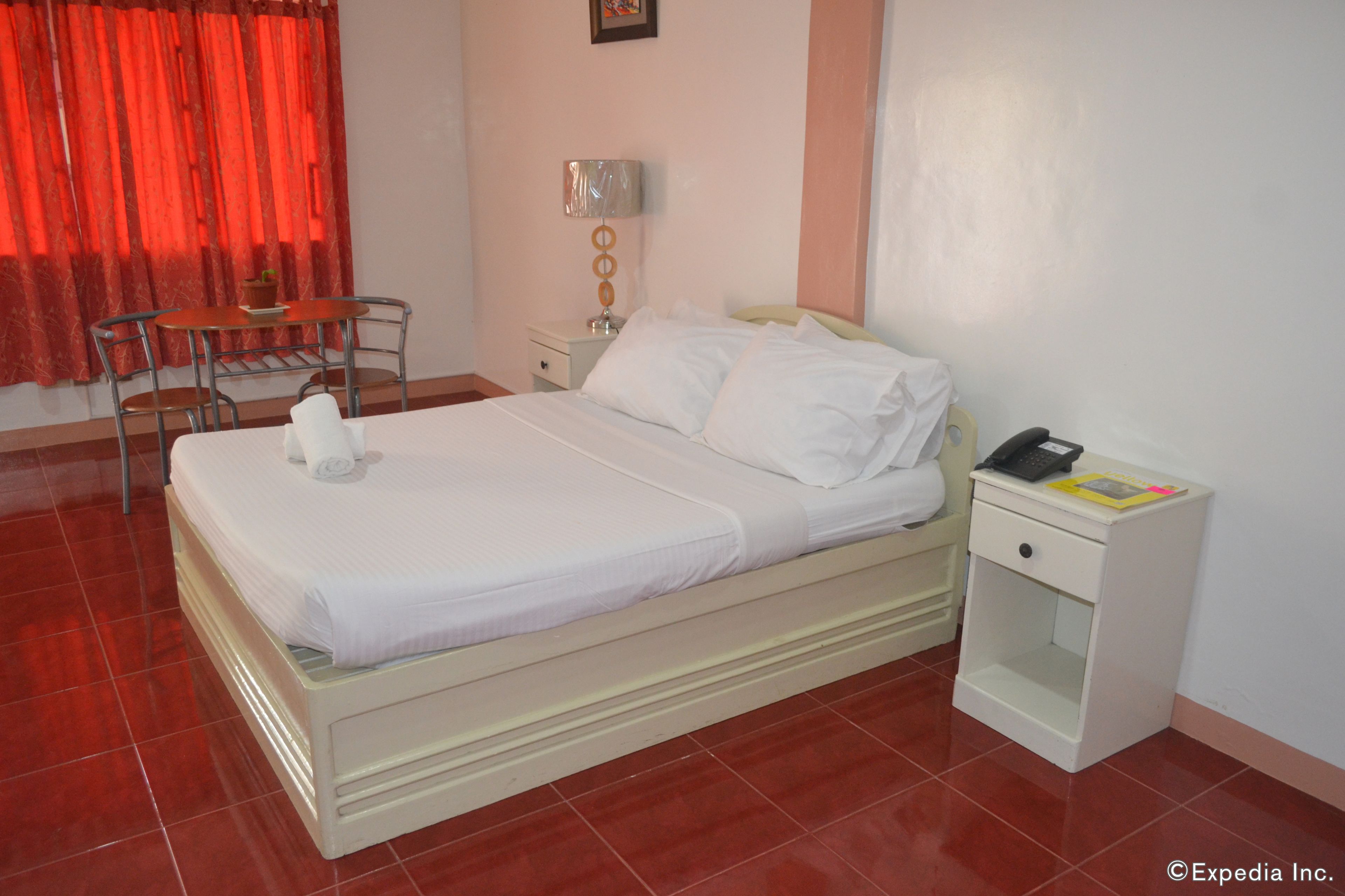 A neatly made bed with white linens, a bedside table with a lamp, and a chair in a room with red curtains and a tiled floor.