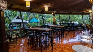 Un área de comedor al aire libre, techada, con mesas y sillas de madera, rodeada de abundante follaje y vistas a una casa tradicional.