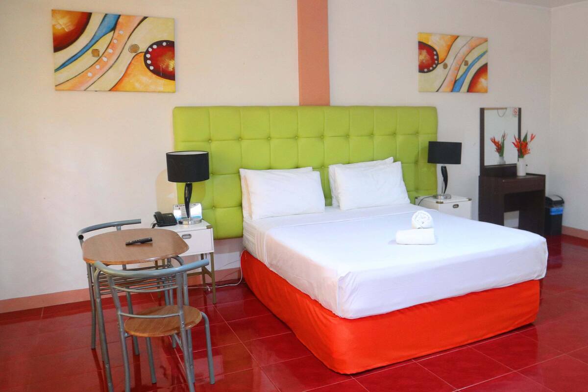 A hotel room with a green upholstered headboard, a bed with white linens, a small table with a chair, a bedside table with a lamp, and artwork on the walls.
