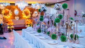 Una mesa de banquete dispuesta con manteles blancos, platos y cristalería, rodeada de arreglos florales y candelabros.