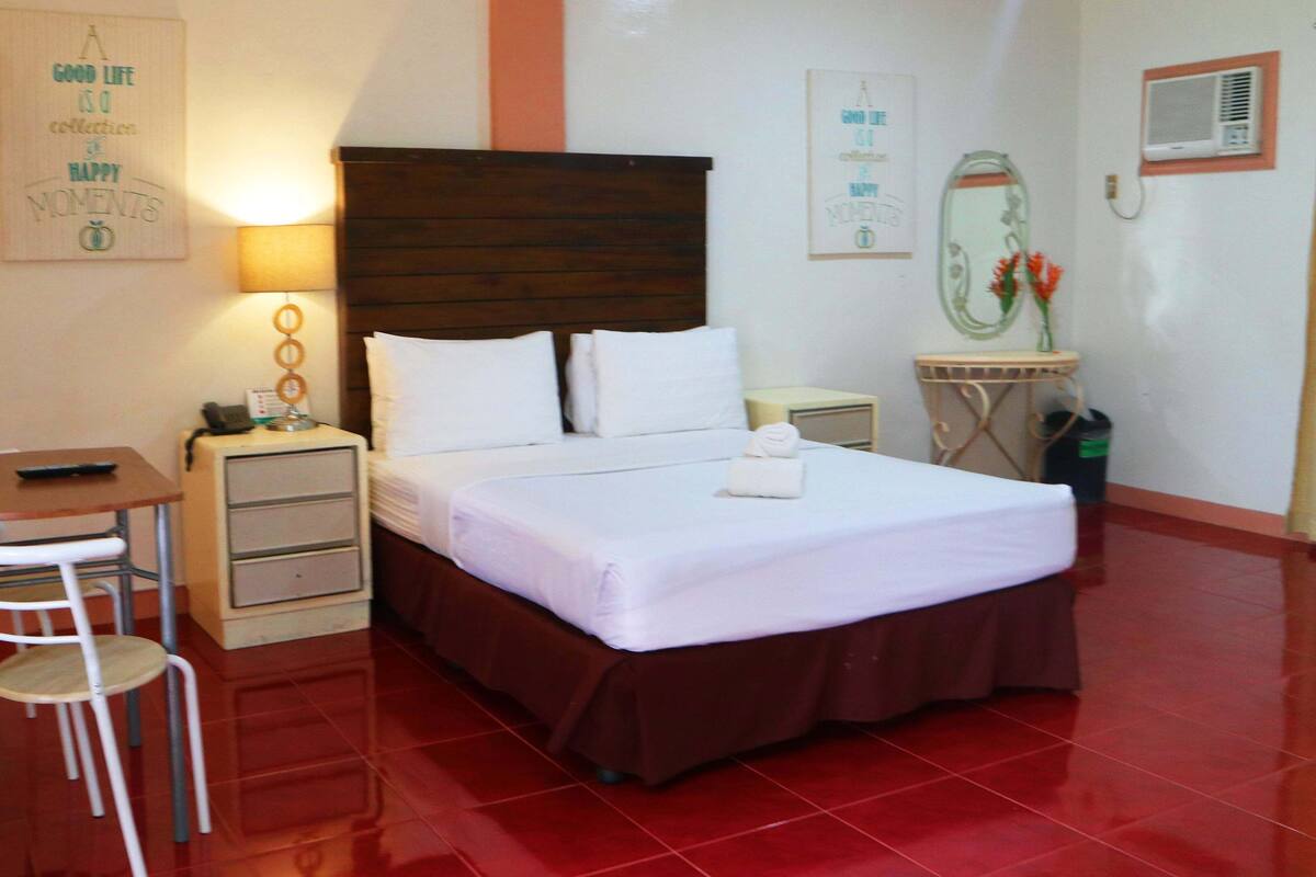 A hotel room with a bed, a desk with a chair, a nightstand, a mirror, and a wall with motivational posters.