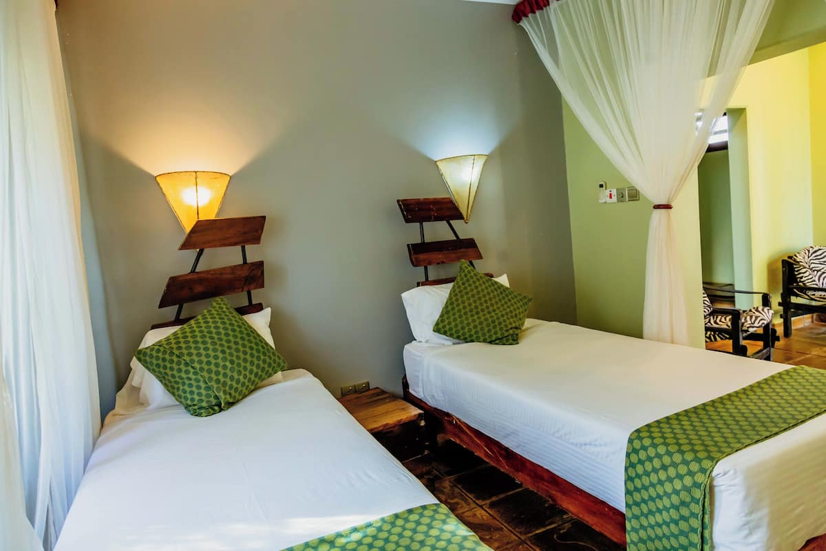 A hotel room with two beds, each with a green patterned bedspread and pillows, a wooden headboard, and a wall-mounted light fixture.