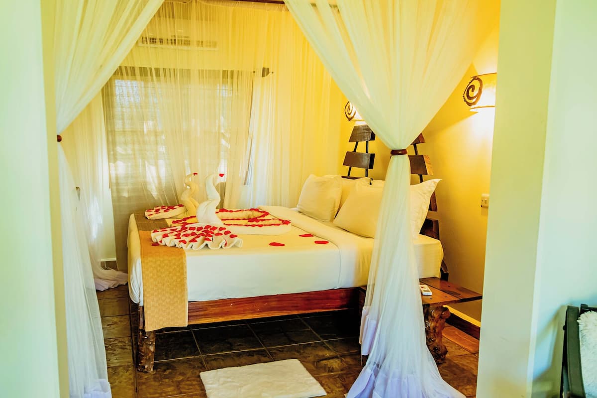 A room with a bed, white curtains, a wooden headboard, a bedside table, and a wall-mounted lamp.