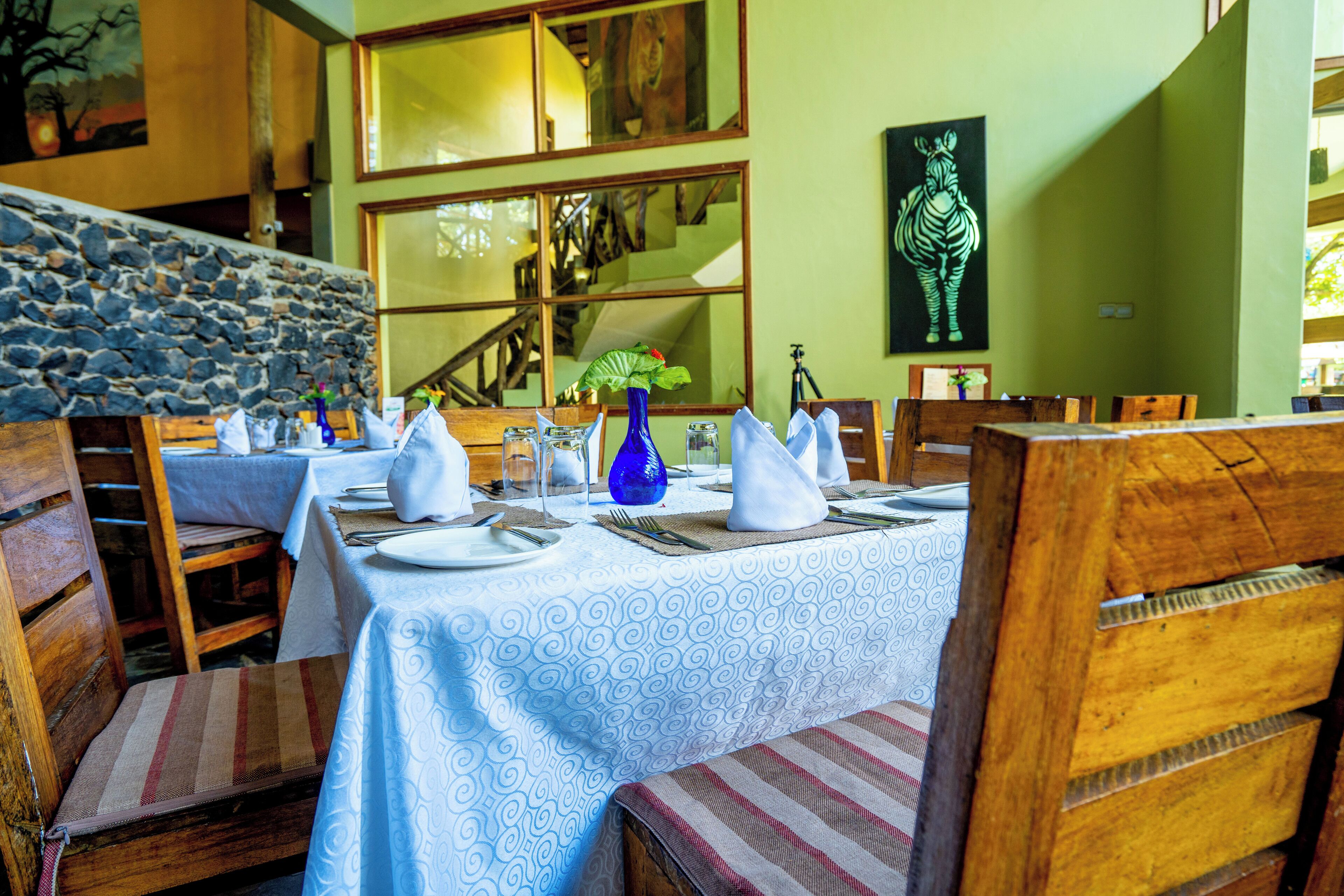 Un comedor con mesas dispuestas para cuatro, sillas de madera, una pared de piedra y una escalera al fondo.