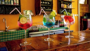 Kolme värikästä cocktail-drinkkiä baaritiskillä, jonka päällä on ruutukuviollinen pöytäliina ja taustalla hyllyllisiä pulloja.
