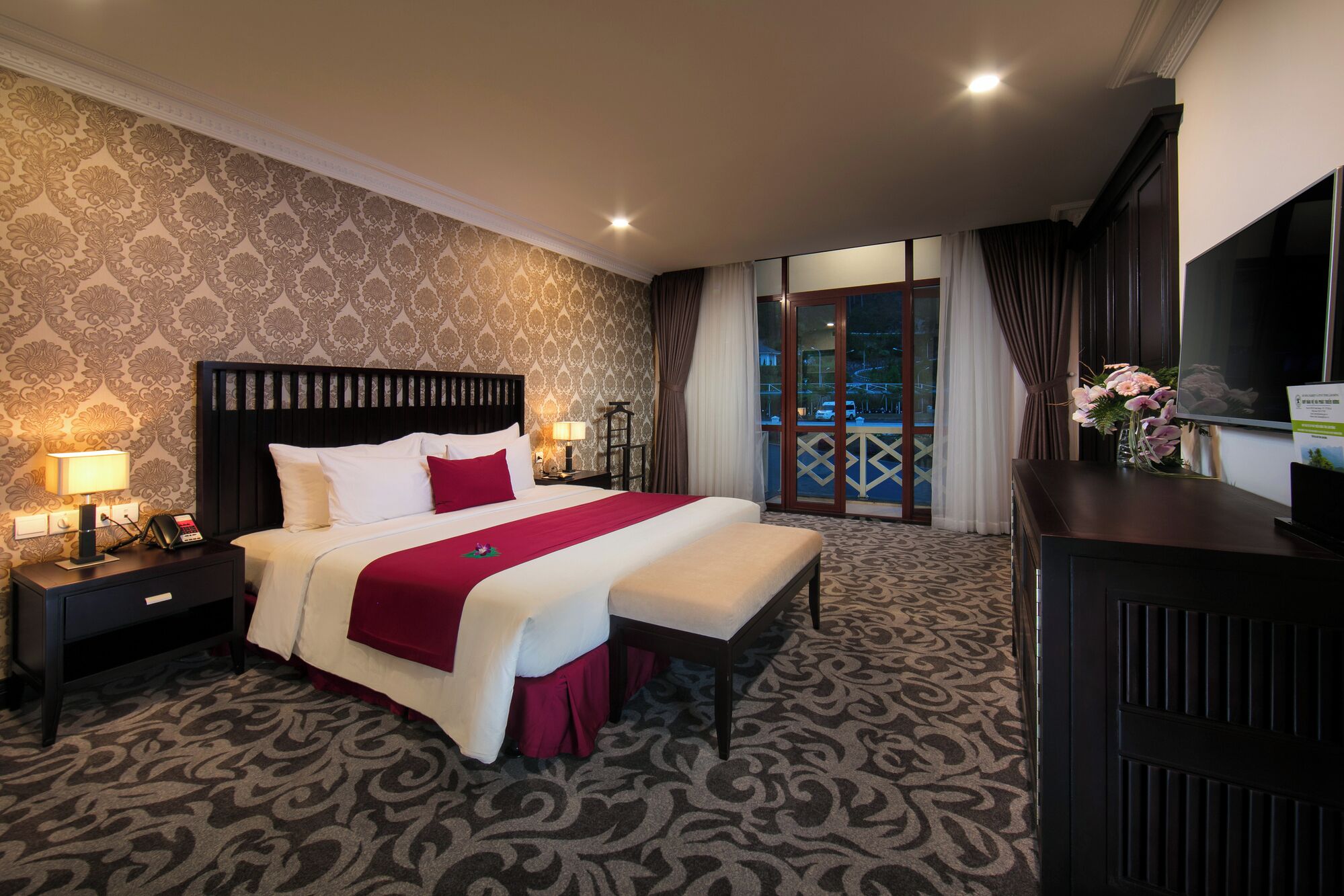 A hotel room with a large bed, a bedside table with a lamp, a TV, and a balcony with curtains.