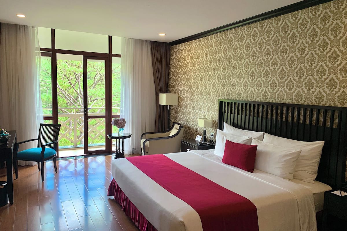 A hotel room with a large bed, a desk, a chair, a small table with a vase, and a view of greenery through the window.