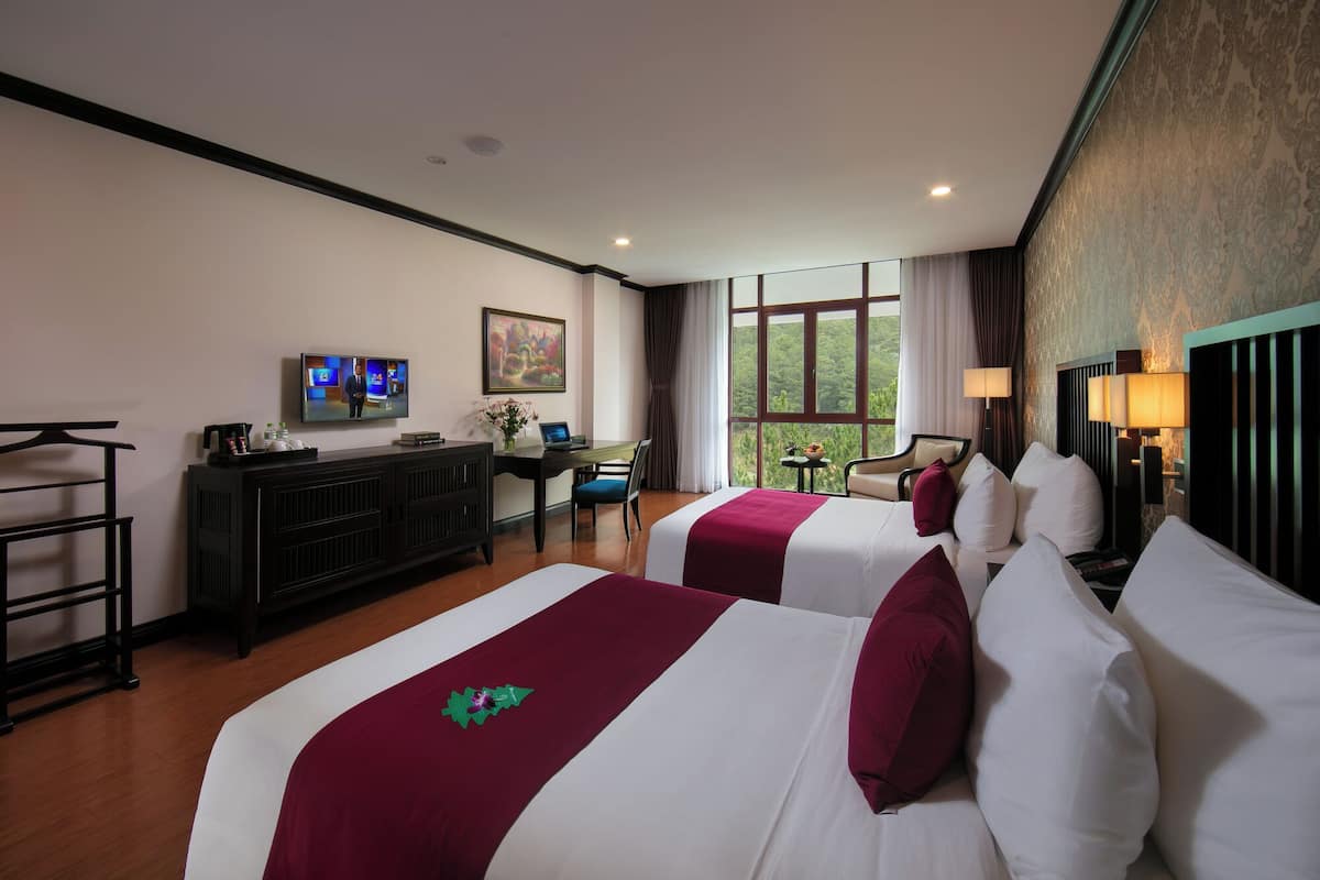 A hotel room with two beds, a desk with a chair, a television, and a window with a view of greenery.