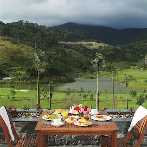 Una mesa dispuesta con alimentos para el desayuno, con vistas a un paisaje pintoresco que incluye un lago y colinas.