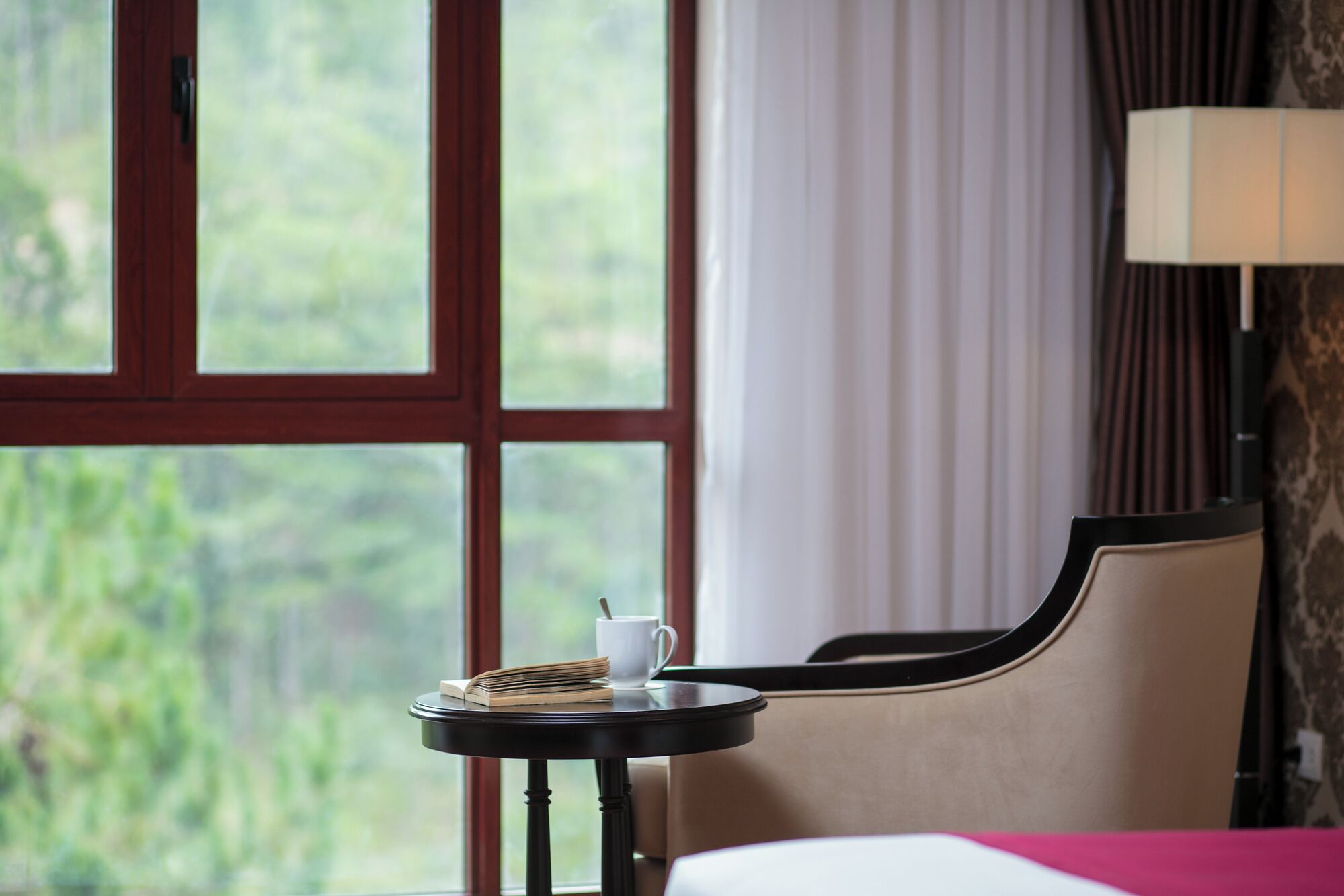 Habitación de hotel con una cama, una mesita con un libro y una taza, una lámpara y una ventana con vistas a la vegetación.