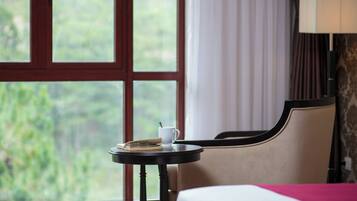 Habitación de hotel con una cama, una mesita con un libro y una taza, una lámpara y una ventana con vistas a la vegetación.