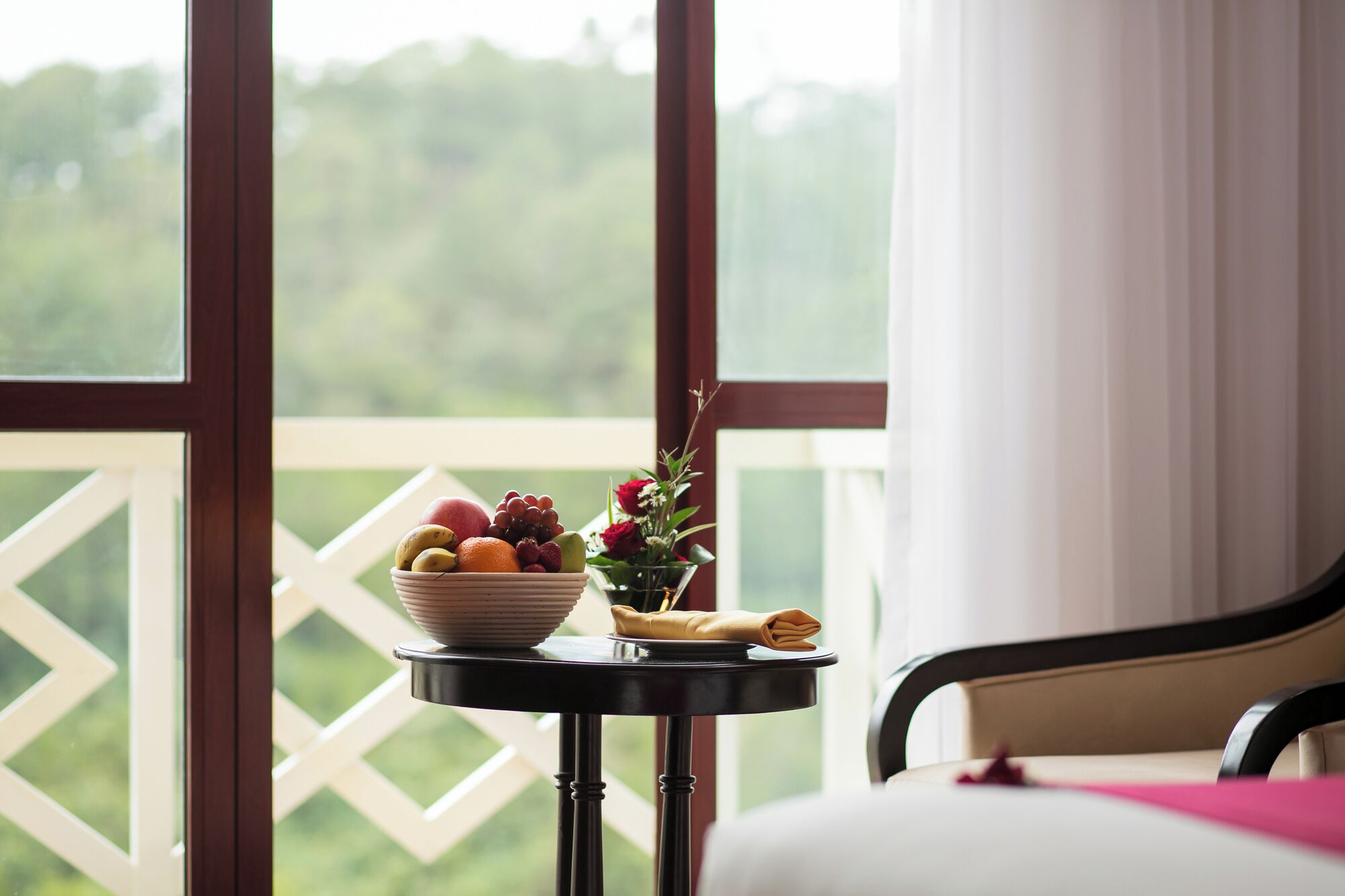 A room with a bed, a small table with a bowl of fruit and a vase of flowers, and a view of greenery through large windows.