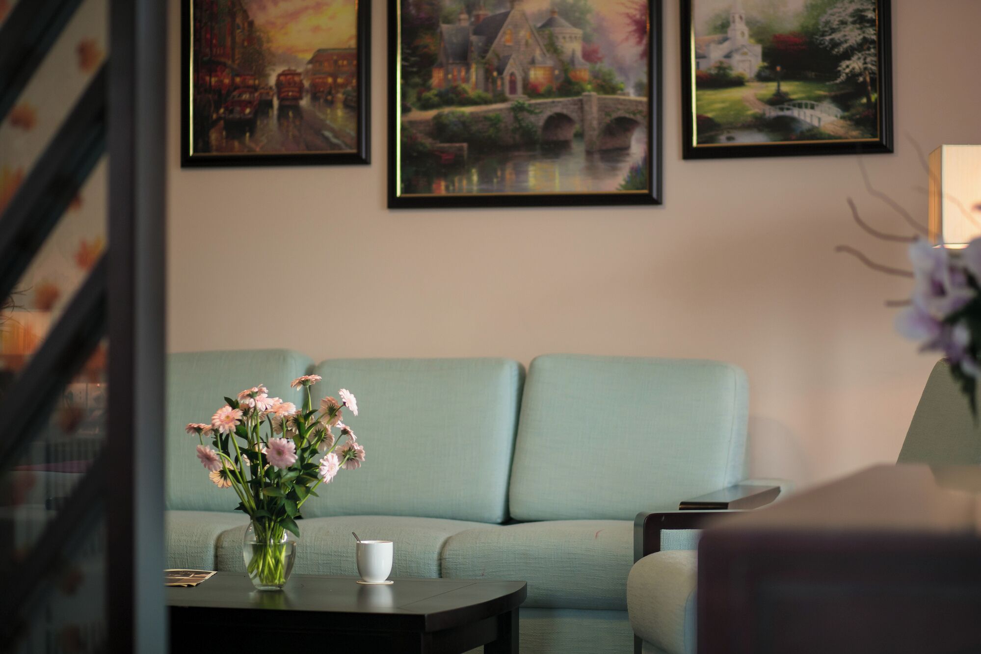 A living room with a light blue sofa, a vase with pink flowers, and a white cup on a table.