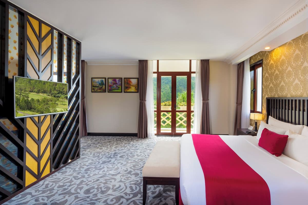 A hotel room with a large bed, a TV, and a balcony with a view of greenery.