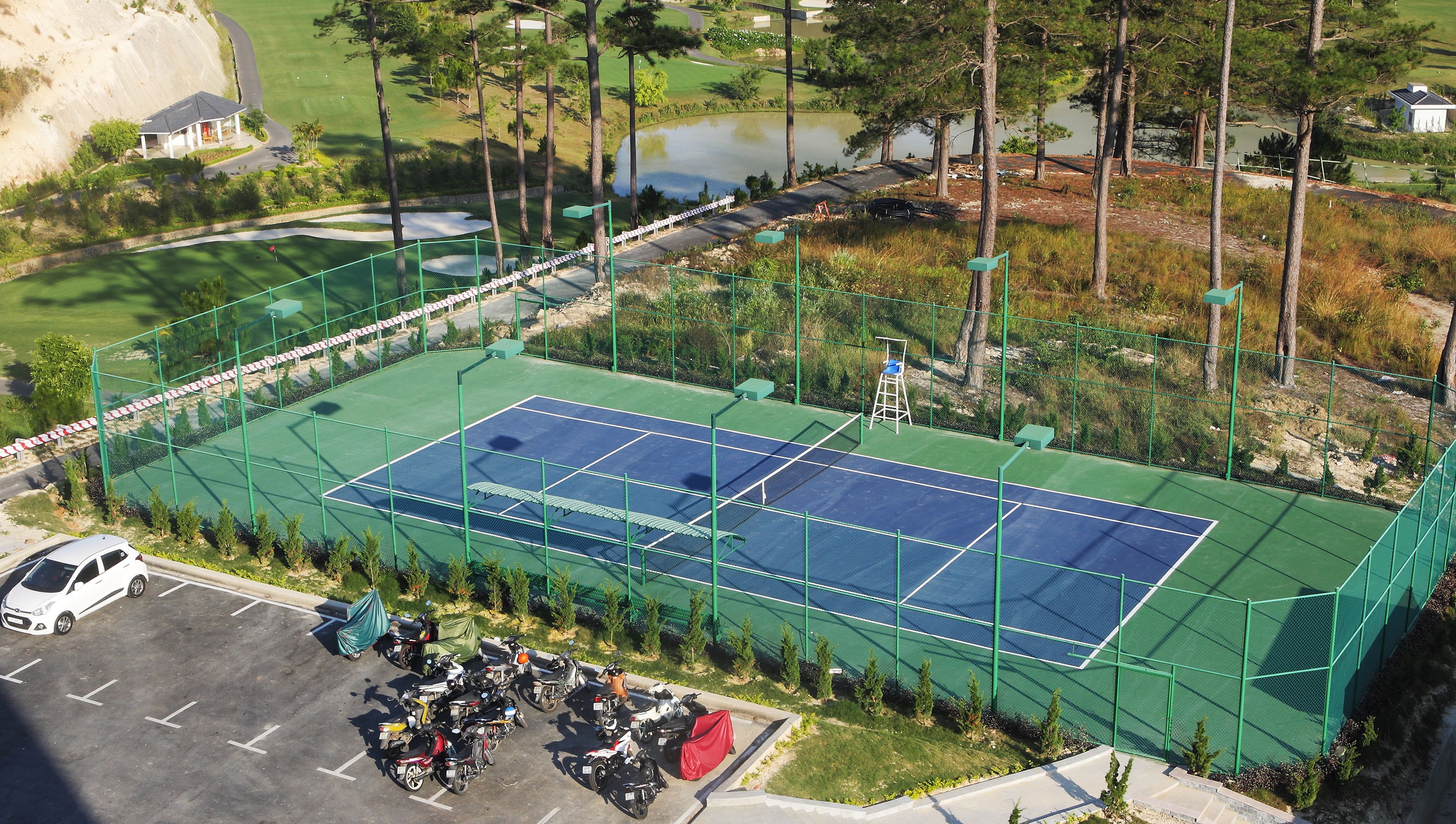 Ein Tennisplatz, umgeben von einem grünen Zaun, mit einem Parkplatz und einigen Bäumen in der Nähe.