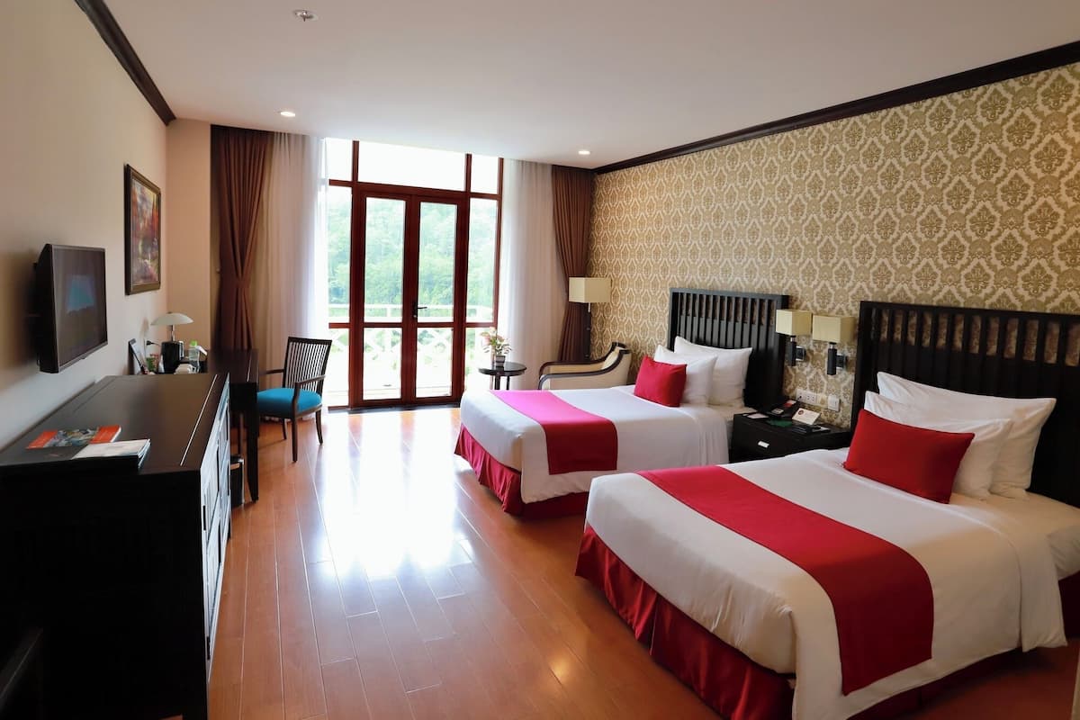 A hotel room with two beds, a desk, a TV, and a view of greenery through the window.