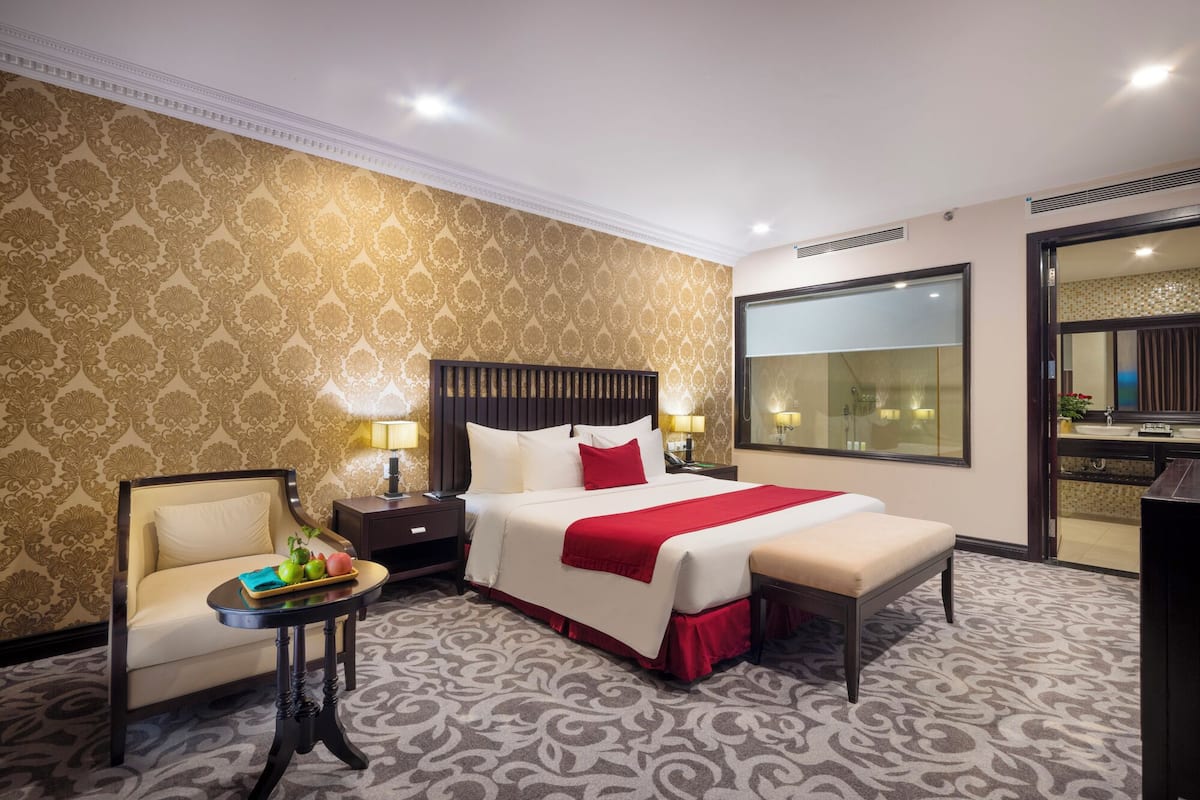 A hotel room with a large bed, a bedside table with a lamp, a chair, a small table with fruit, and a large mirror.