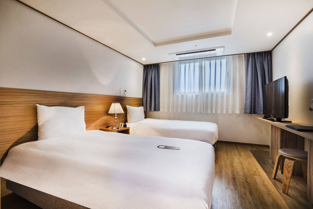 A hotel room with two beds, a TV, a desk, and a window with curtains.