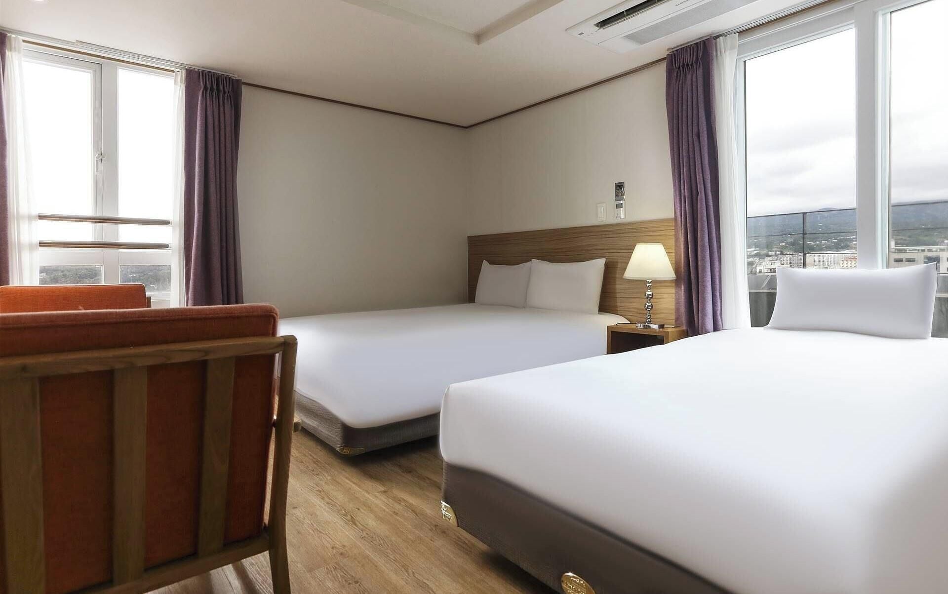 A hotel room with two beds, a wooden chair, a nightstand with a lamp, and large windows offering a city view.