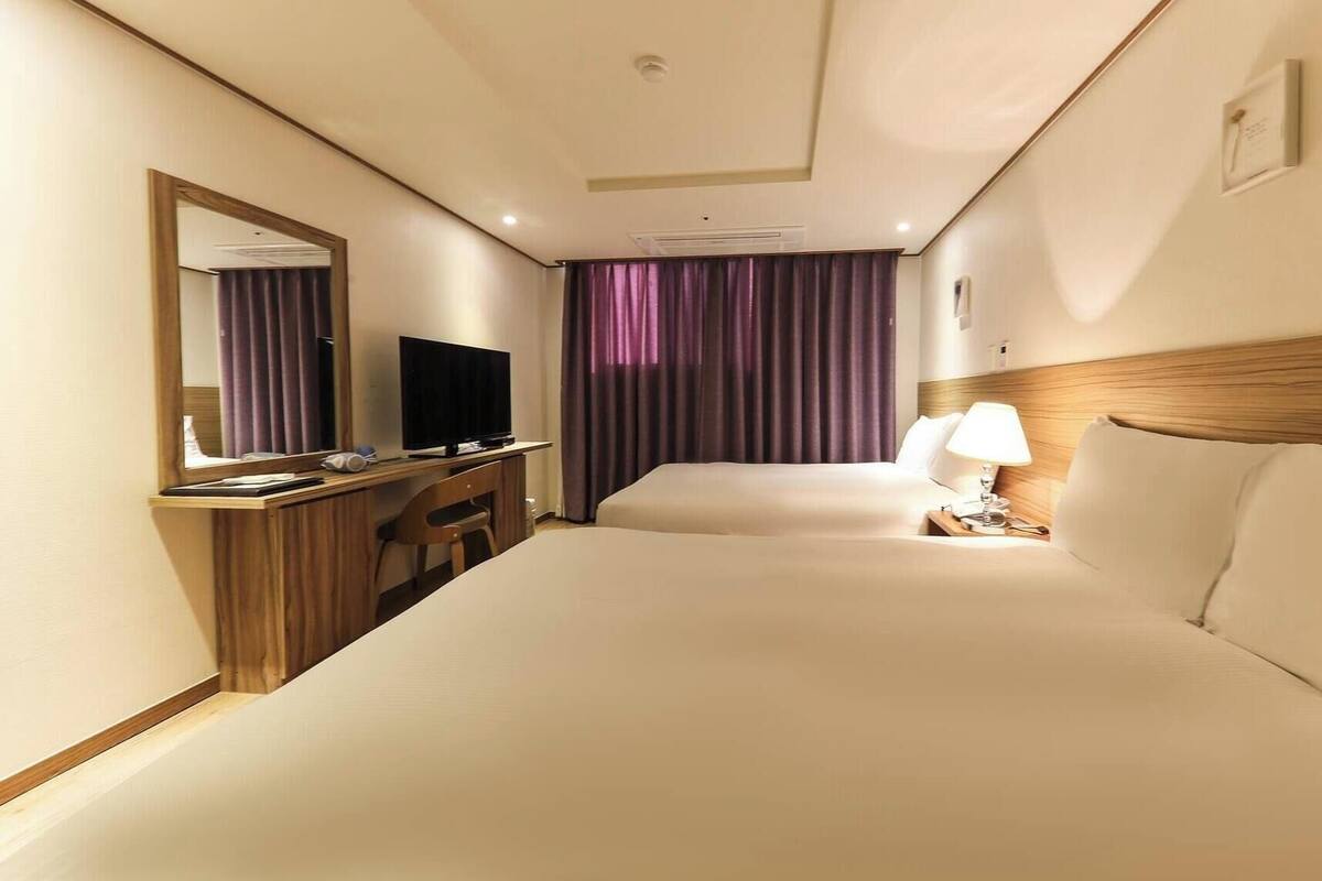A hotel room with a bed, a desk with a television, a chair, a nightstand with a lamp, and a window with purple curtains.