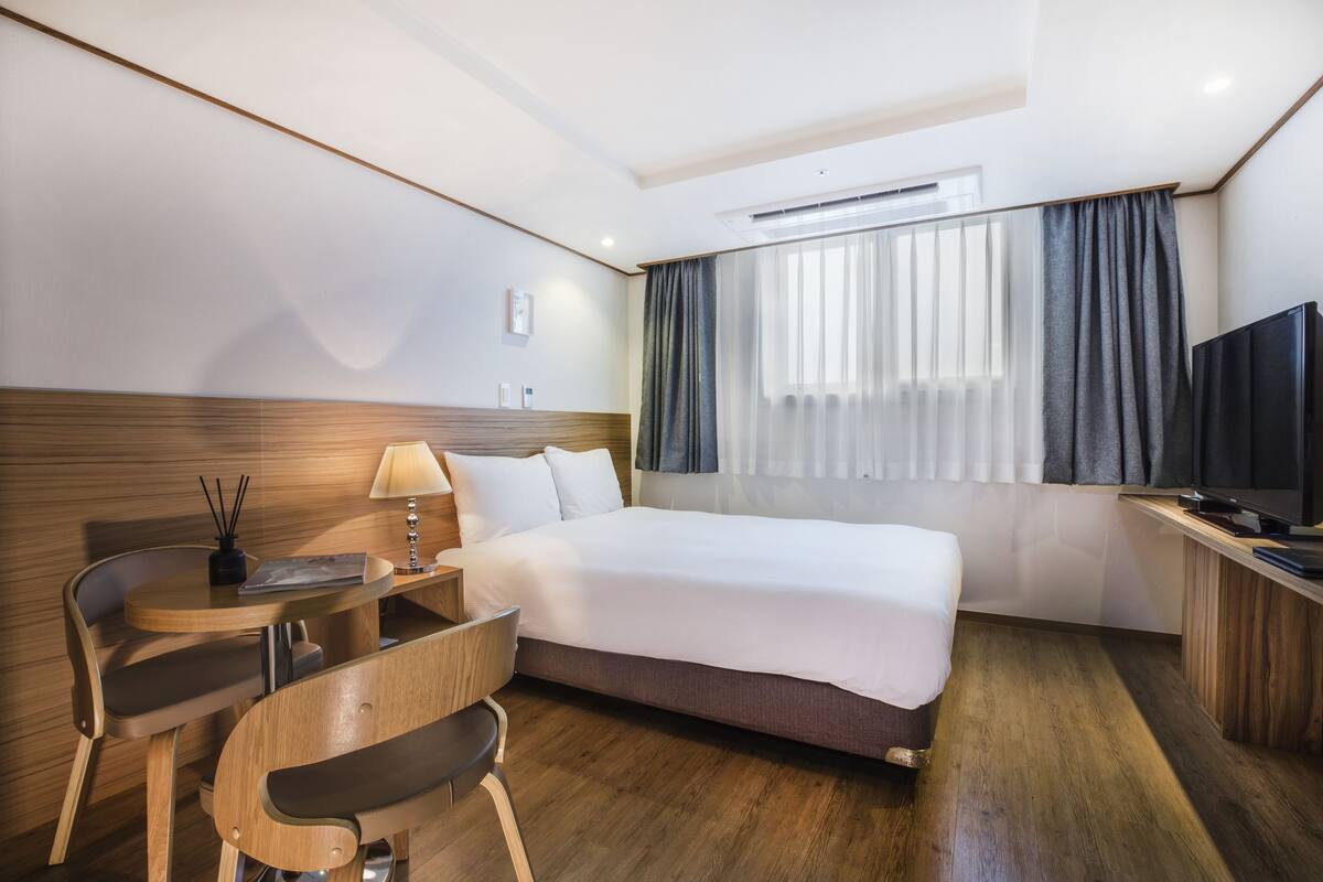A hotel room with a bed, a desk, a chair, a TV, and a window with curtains.