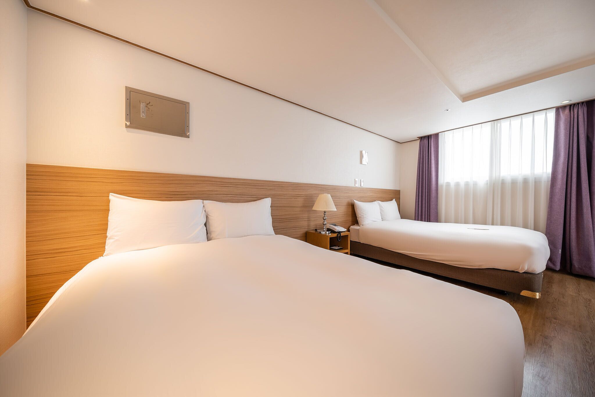 Habitación de hotel con dos camas, cabecero de madera, mesita de noche con lámpara y ventana con cortinas.