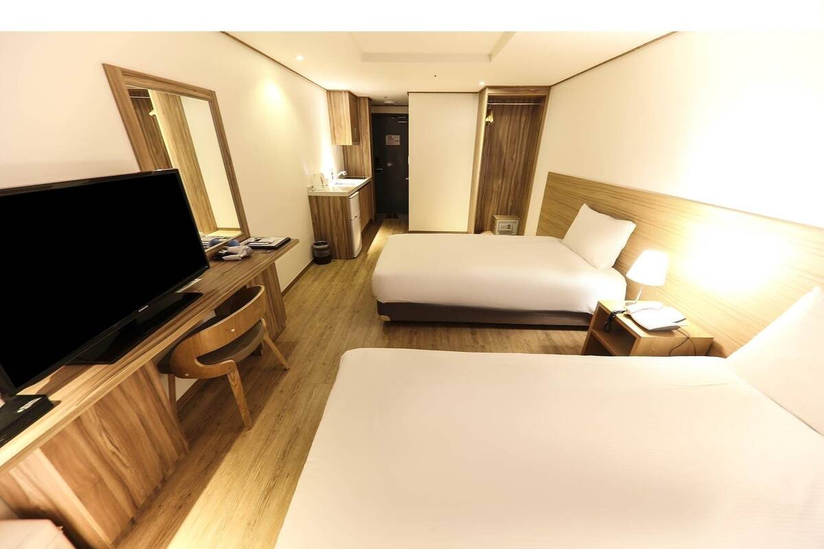 A hotel room with a bed, a desk with a TV, a bedside table with a lamp, and a bathroom with a sink and mirror.