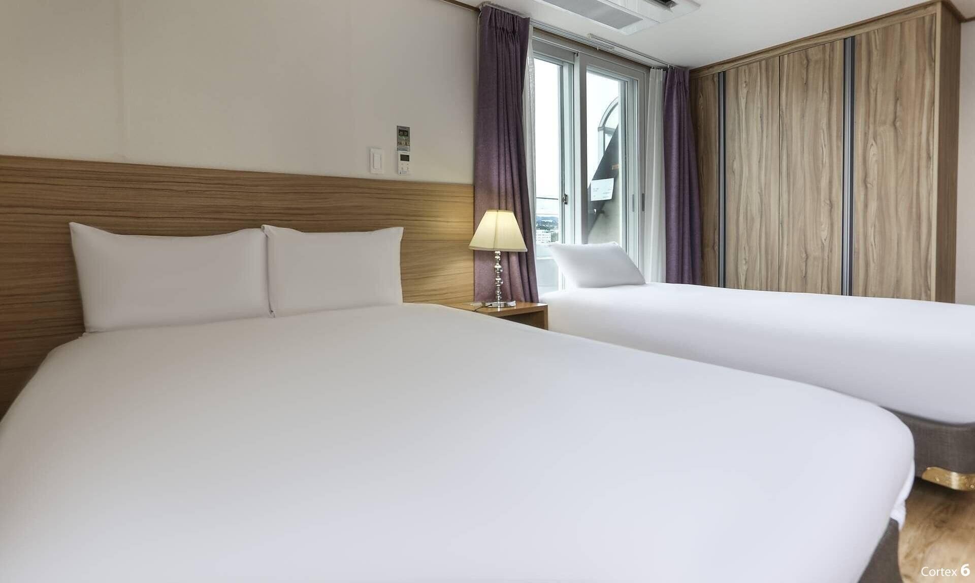 A hotel room with two beds, a window with curtains, a wooden headboard, and a wall-mounted air conditioning unit.