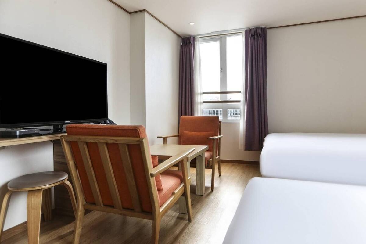 A hotel room with a flat-screen TV, a desk, a chair, a bed, and a window with curtains.