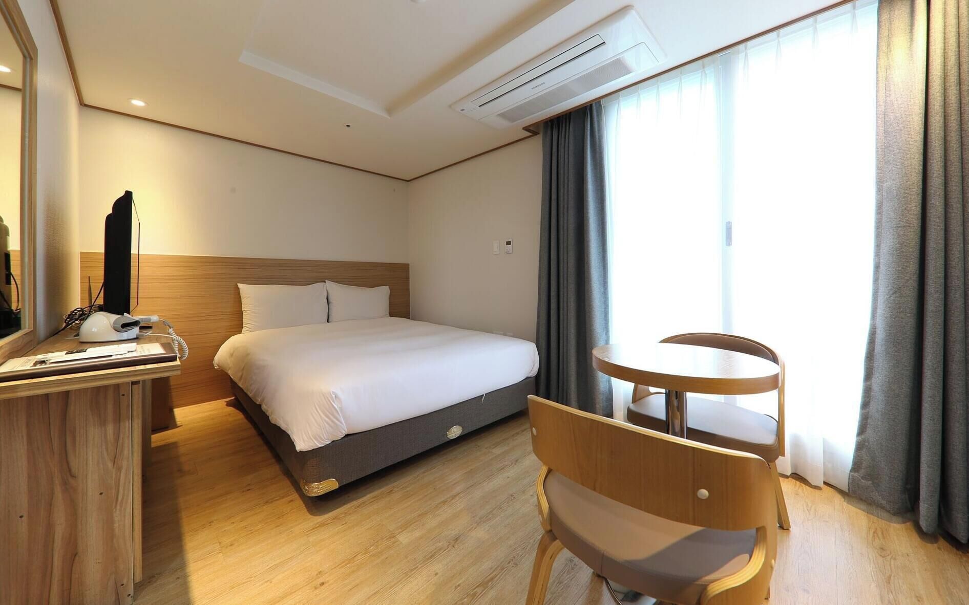 Een hotelkamer met een bed, een bureau met telefoon en oplader, een stoel en een raam met gordijnen.