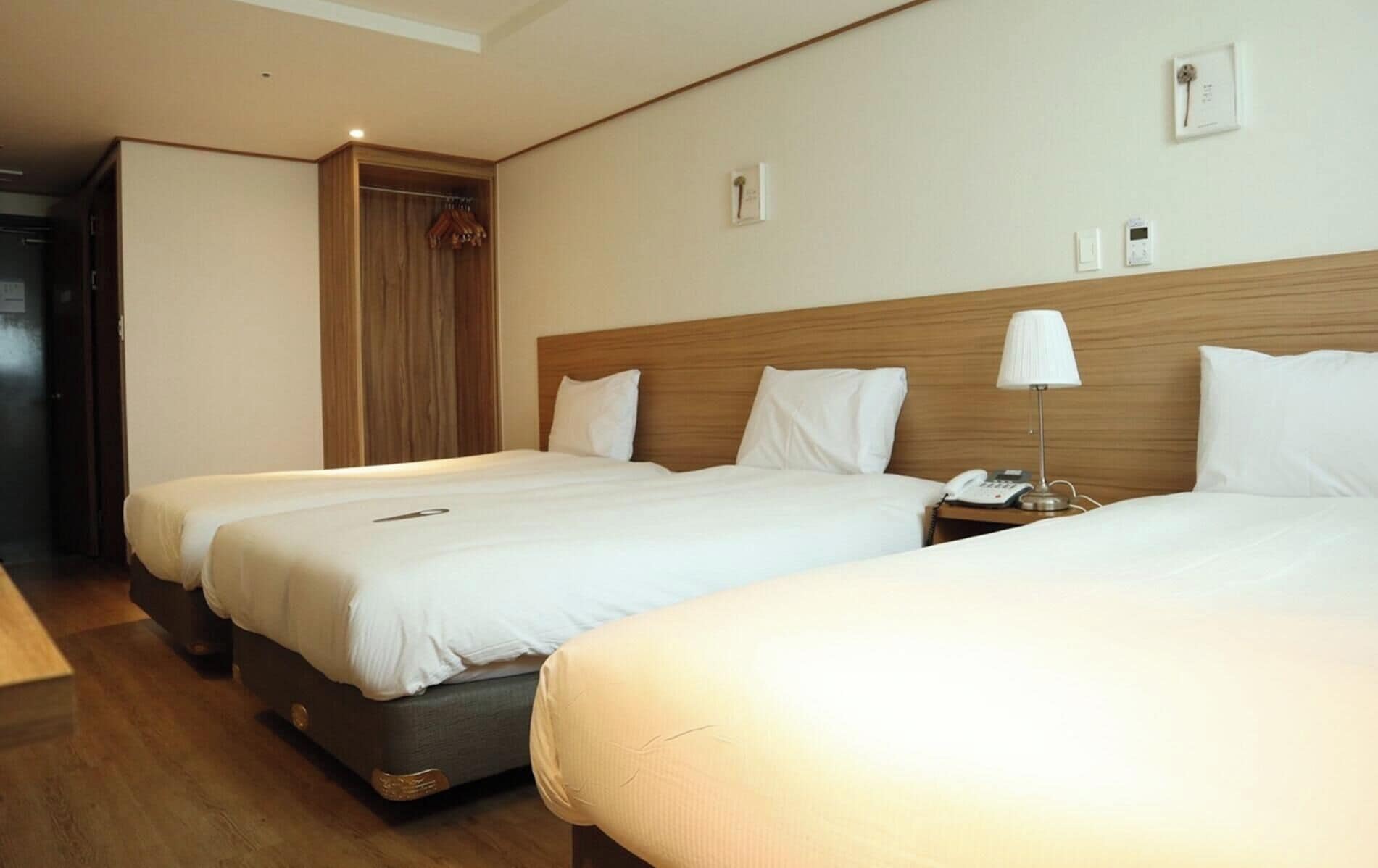 Une chambre d’hôtel avec deux lits simples, une tête de lit en bois, une table de chevet avec une lampe et une armoire.