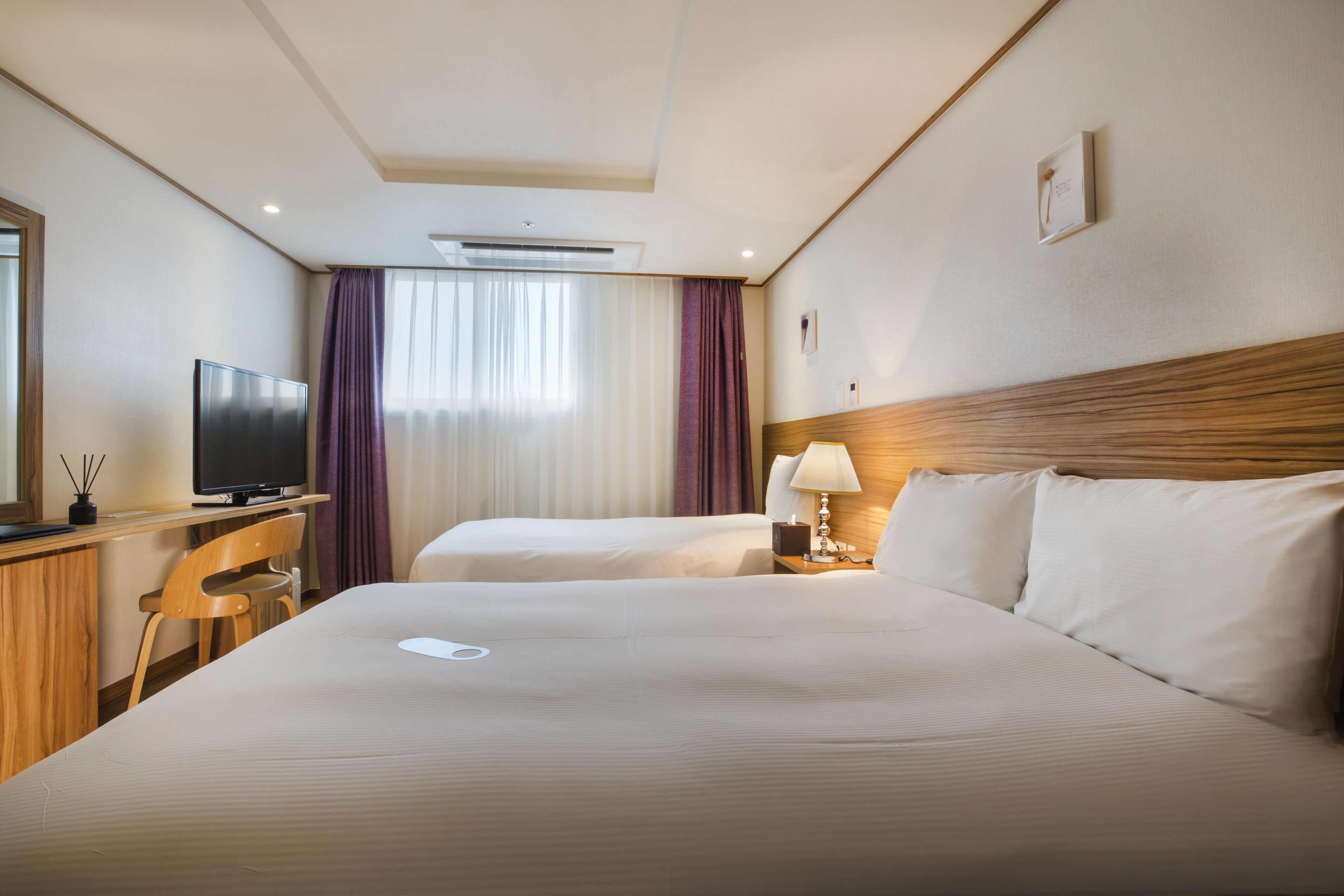 Ein Hotelzimmer mit zwei Betten, einem Schreibtisch, einem Fernseher, einer Lampe und einem Fenster mit Vorhängen.