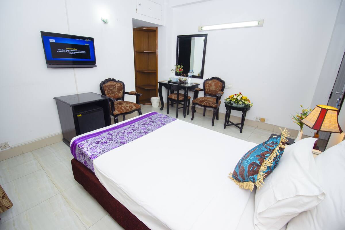 A hotel room with a bed, a desk with two chairs, a television, a wardrobe, and a small table with flowers.
