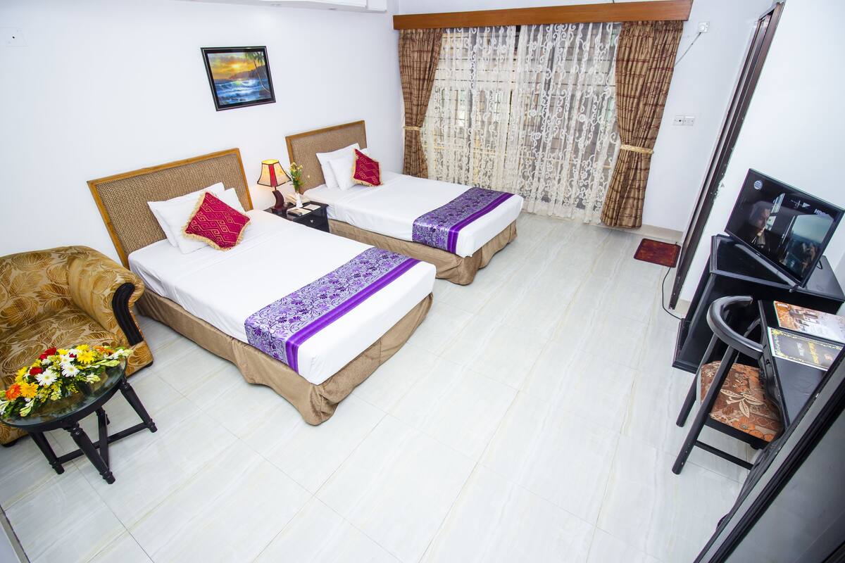 A hotel room with two beds, a sofa, a small table with flowers, a TV, and curtains.
