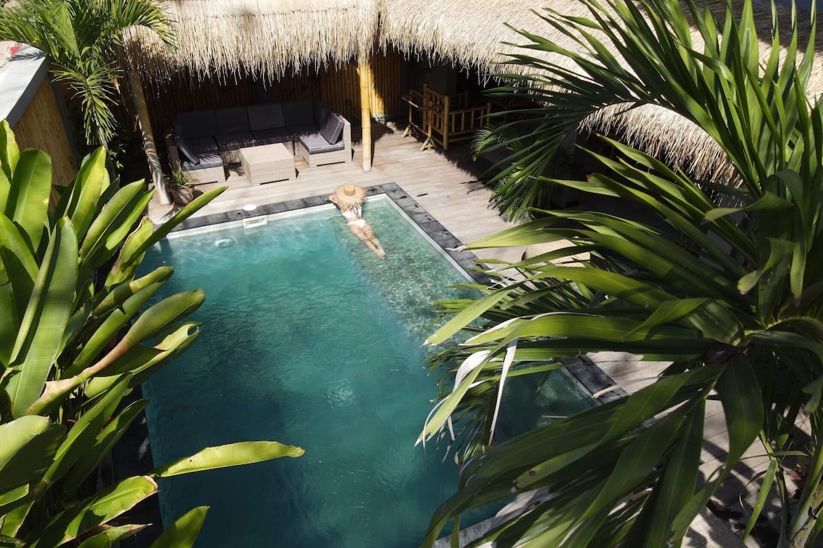 A swimming pool surrounded by tropical plants, with a thatched roof structure and outdoor seating area.