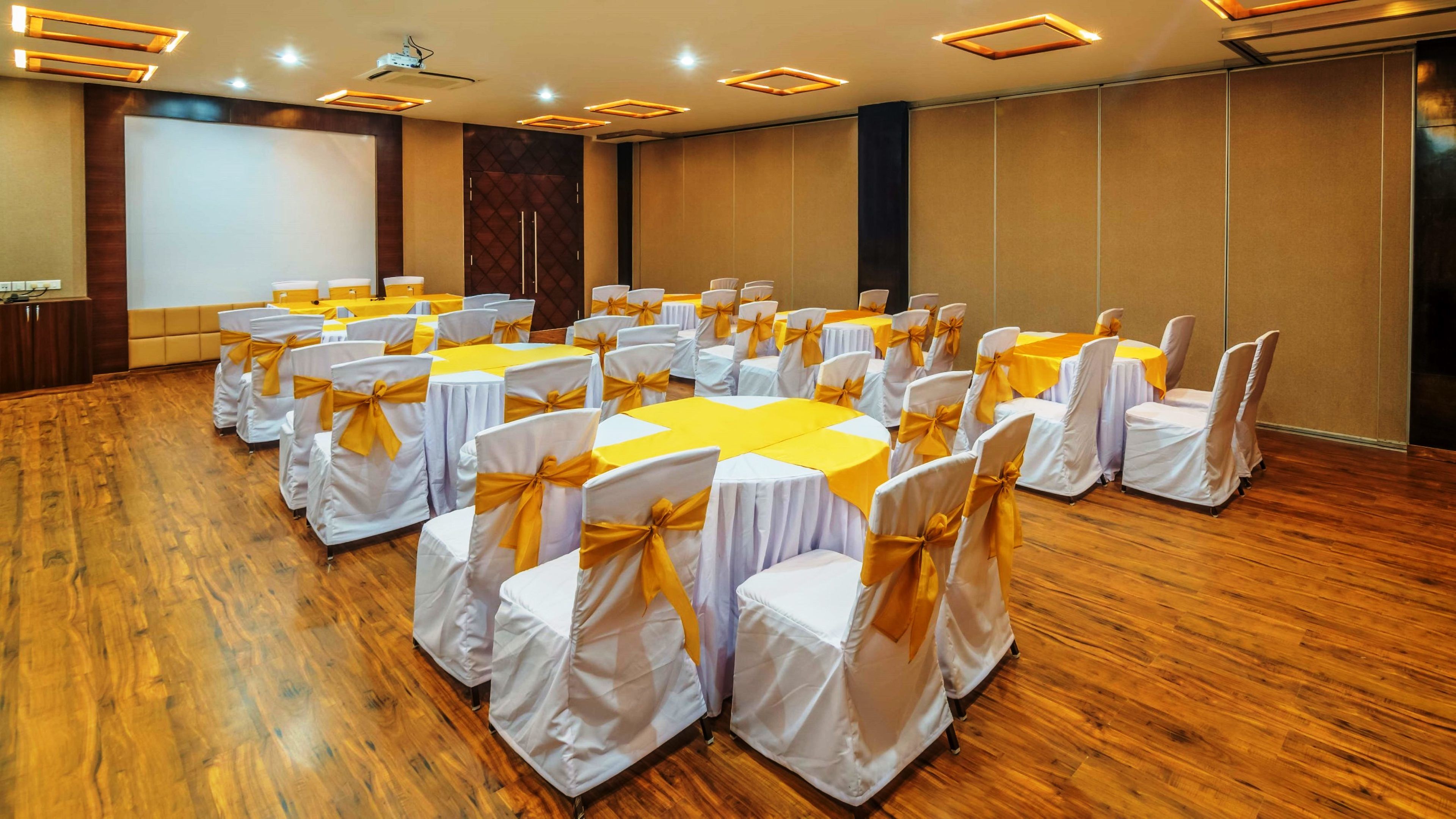 A conference room with round tables and chairs, wooden flooring, and a projector screen.
