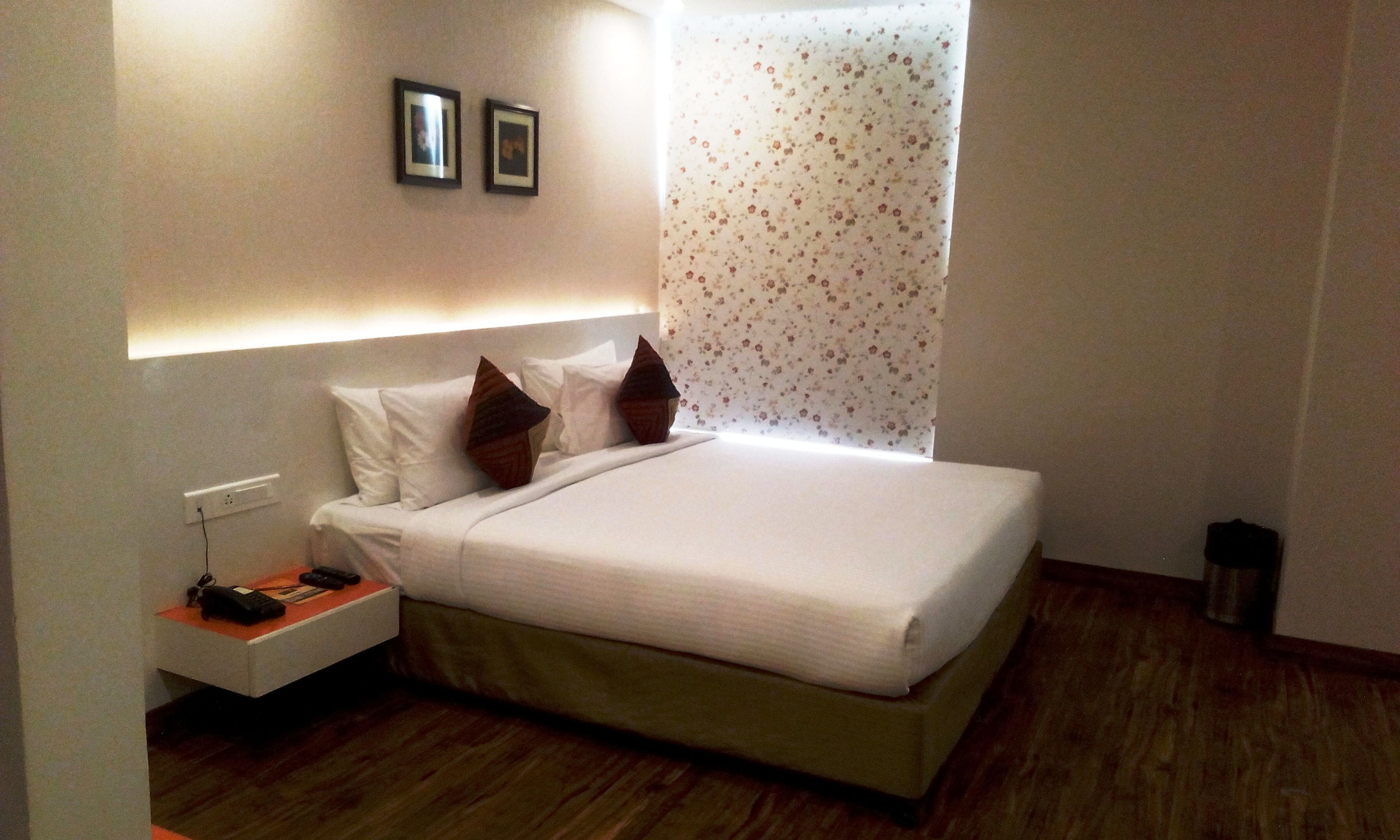 A neatly made bed with white linens and two decorative pillows, a bedside table with a remote control and a phone, and a wall-mounted light fixture.