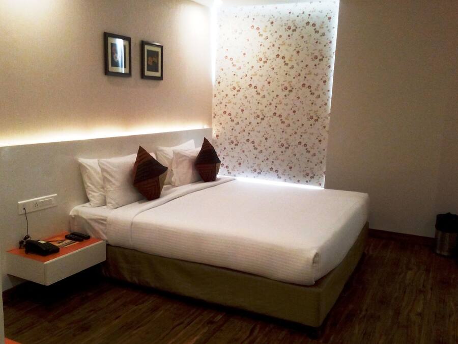A neatly made bed with white linens and two decorative pillows, a bedside table with a remote control and a phone, and a wall-mounted light fixture.