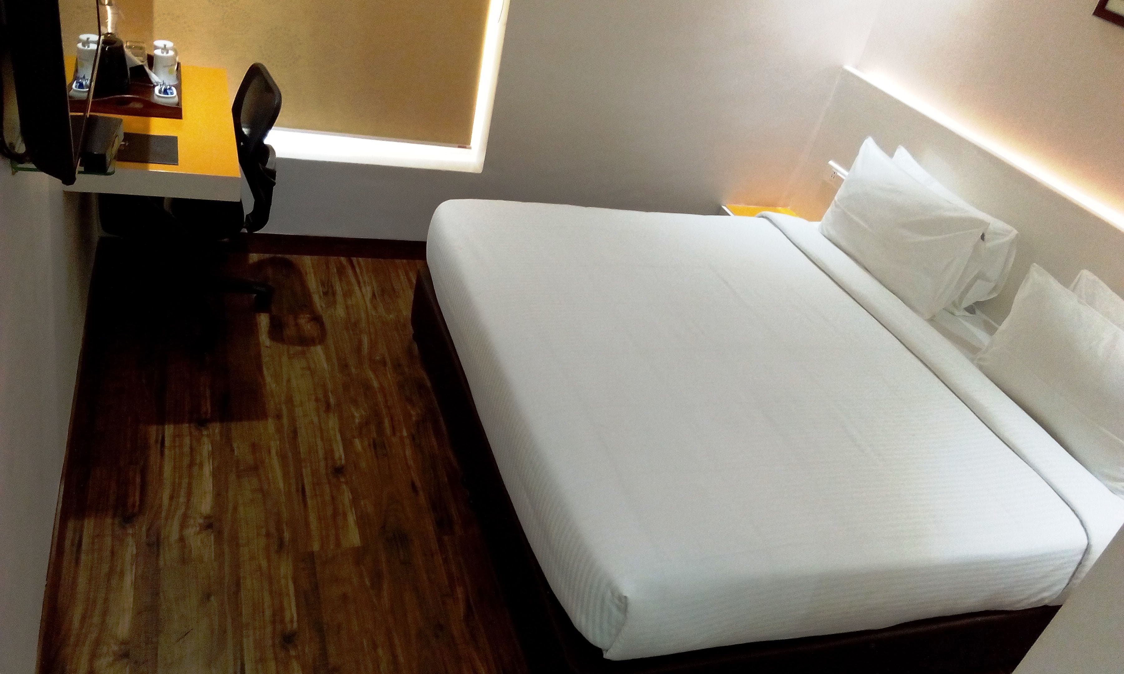 Habitación de hotel con piso de madera, una cama con ropa blanca, un escritorio con silla y una ventana que proporciona luz natural.