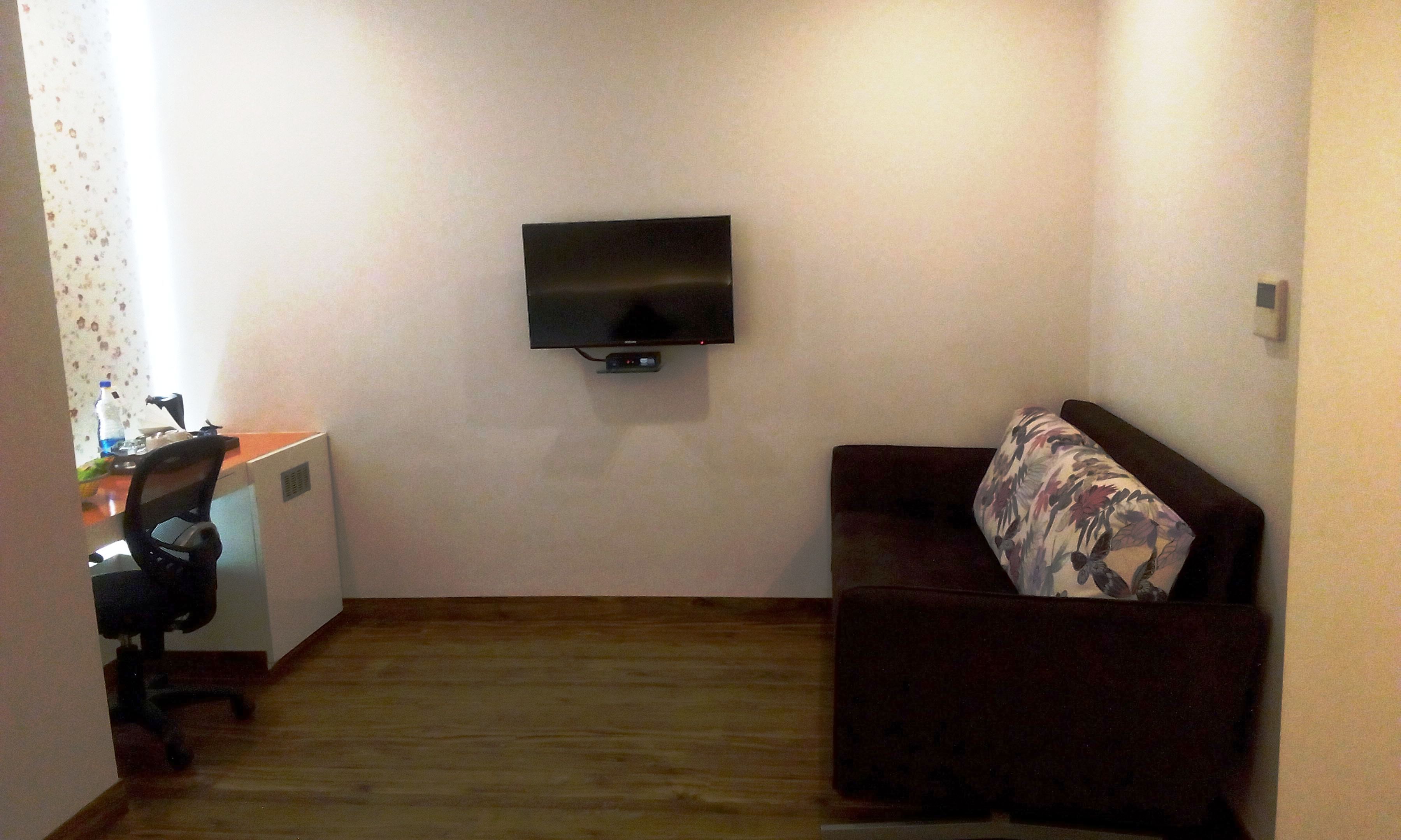 Une petite pièce avec un plancher en bois, un canapé brun foncé agrémenté d’un coussin à motifs floraux, un téléviseur à écran plat fixé au mur et un bureau avec une chaise.