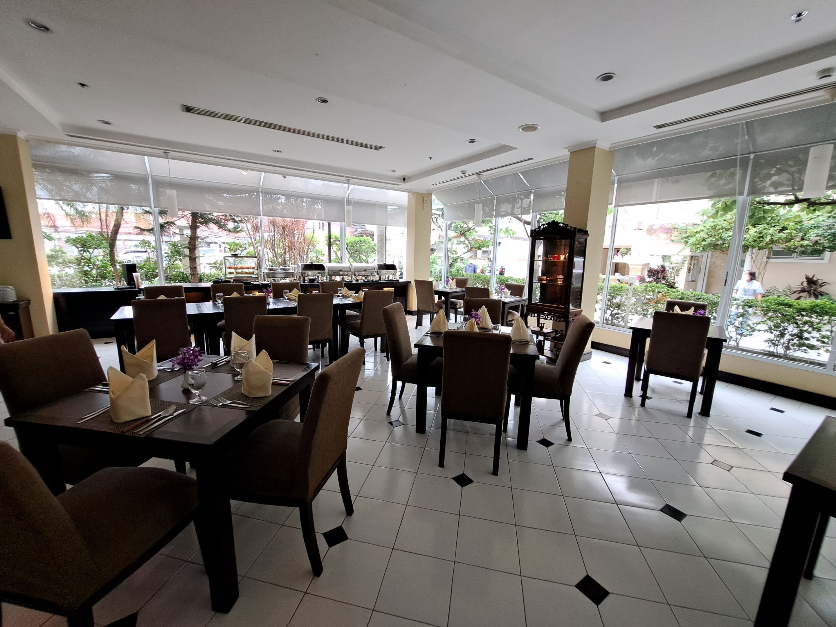 Un restaurante espacioso con techos altos, ventanas amplias y piso de baldosas. Las mesas están dispuestas con sillas y servilletas.