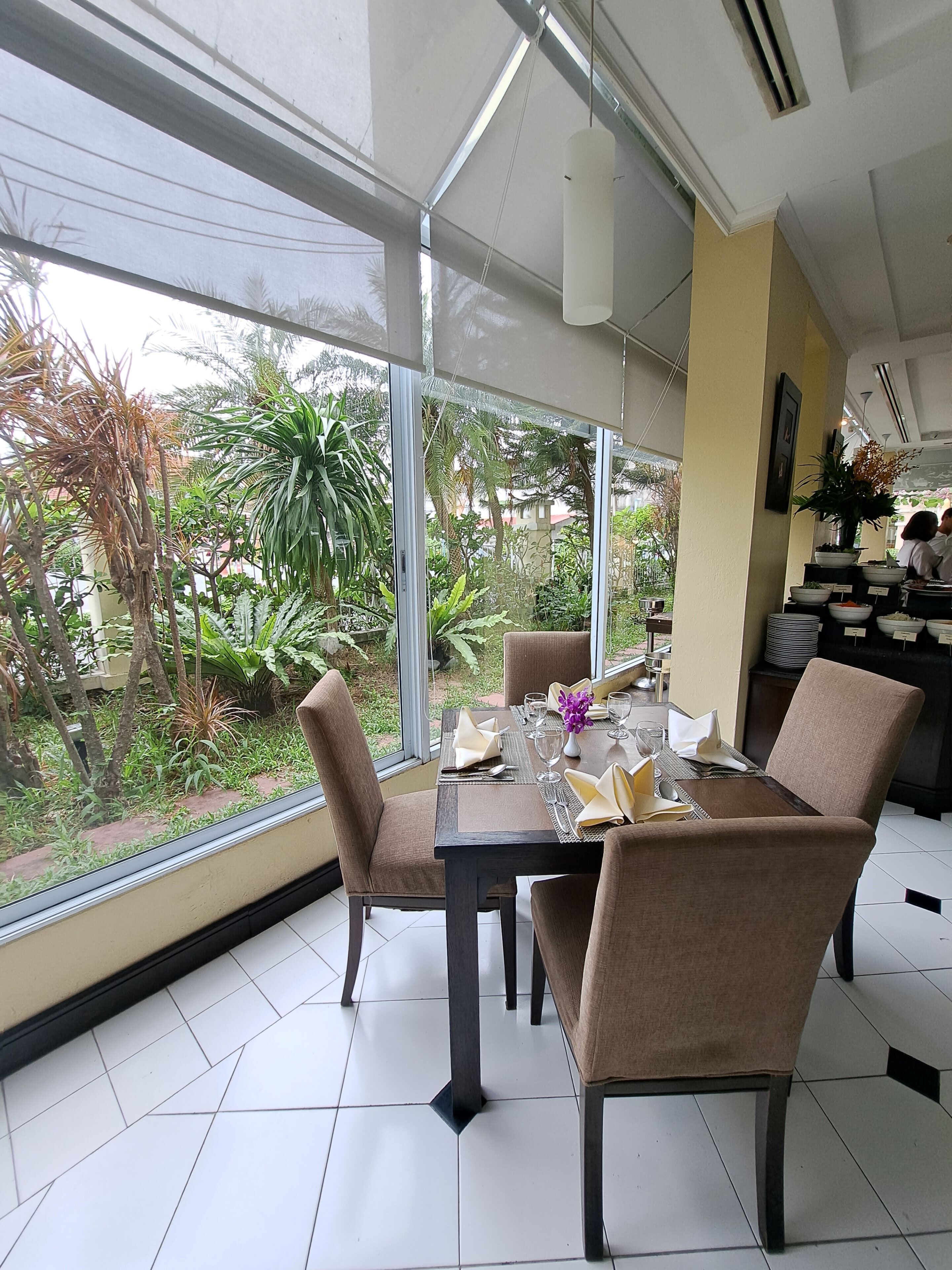 A dining area with a table set for four, surrounded by chairs, large windows, and a view of greenery outside.