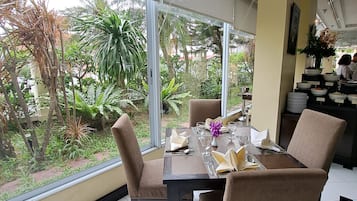 A dining area with a table set for four, surrounded by chairs, large windows, and a view of greenery outside.