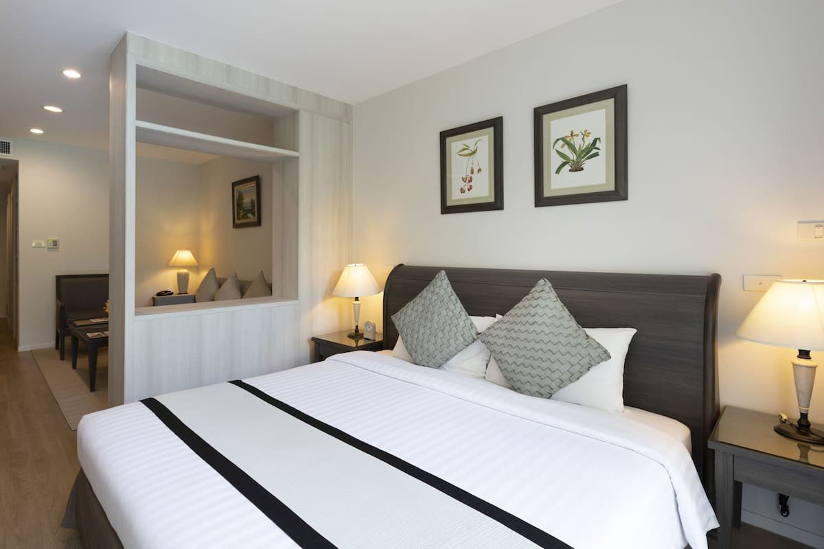 A hotel room with a large bed, two bedside tables with lamps, a nightstand, a chair, a desk, a mirror, and two framed botanical illustrations on the wall.