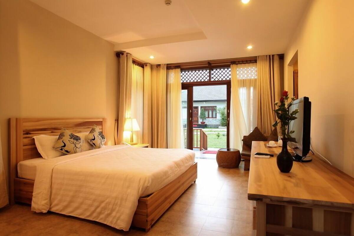 A hotel room with a bed, bedside table, lamp, and a view of the outside.